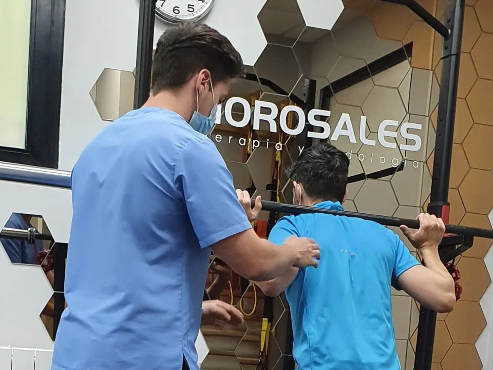 Un fisioterapeuta de Fisiorosales guía a un paciente, de espaldas, en una sentadilla con barra, delante de la pared con el logo del centro